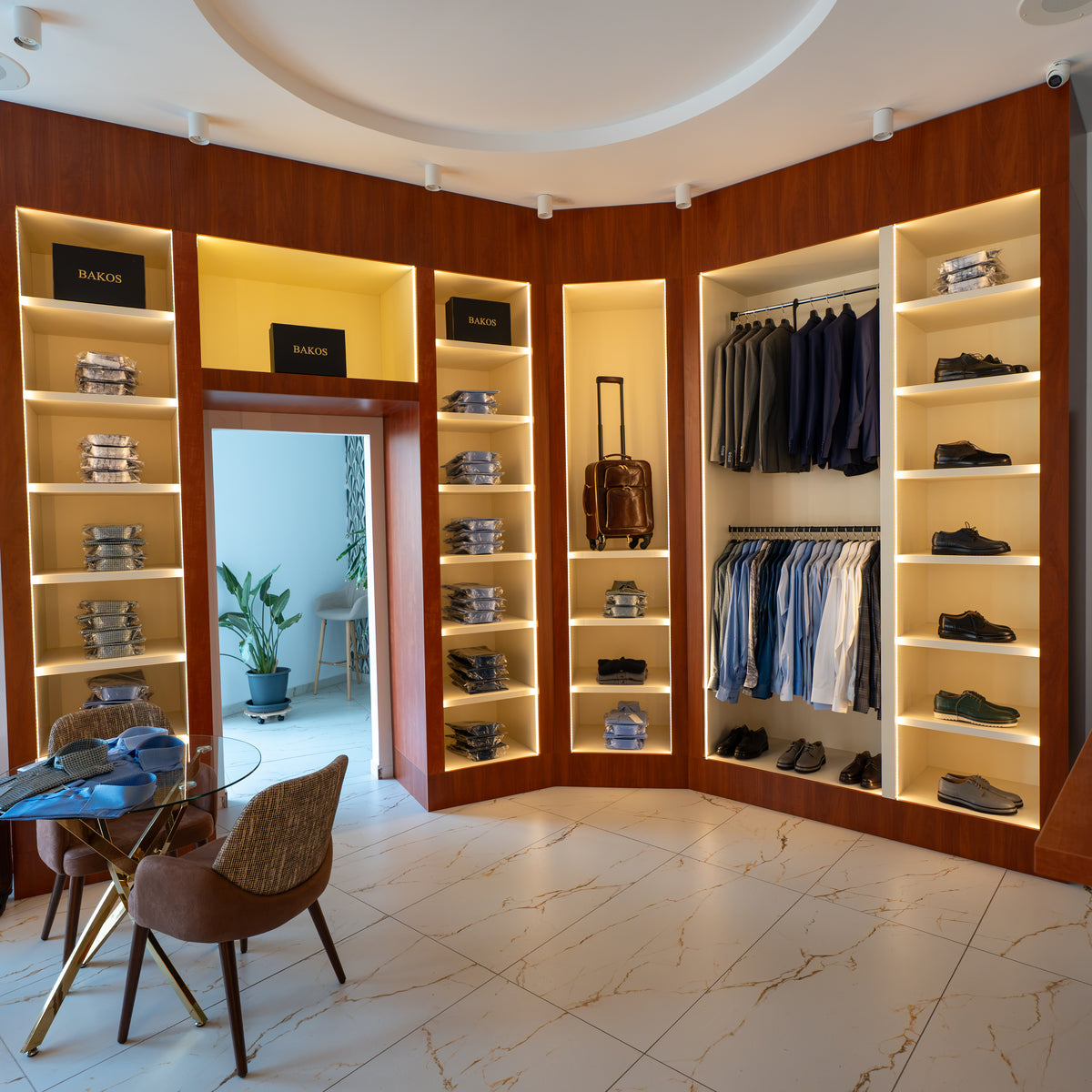 A luxury men's clothing showroom with warm wood-paneled walls, built-in shelving, and lighting. Shelves display stacks of folded shirts and shoes, while hanging racks display suits and dress shirts.