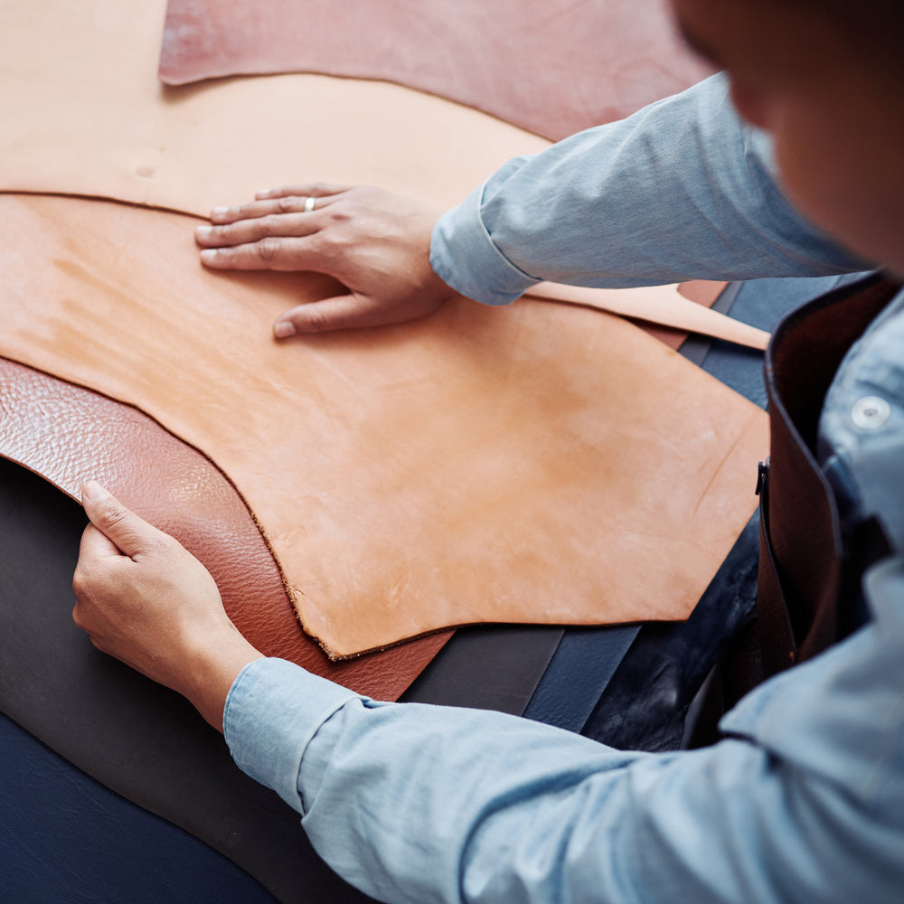 A close-up of a person's hands smoothing out a large tan leather sheet, with other colored leather sheets stacked underneath on a worktable.